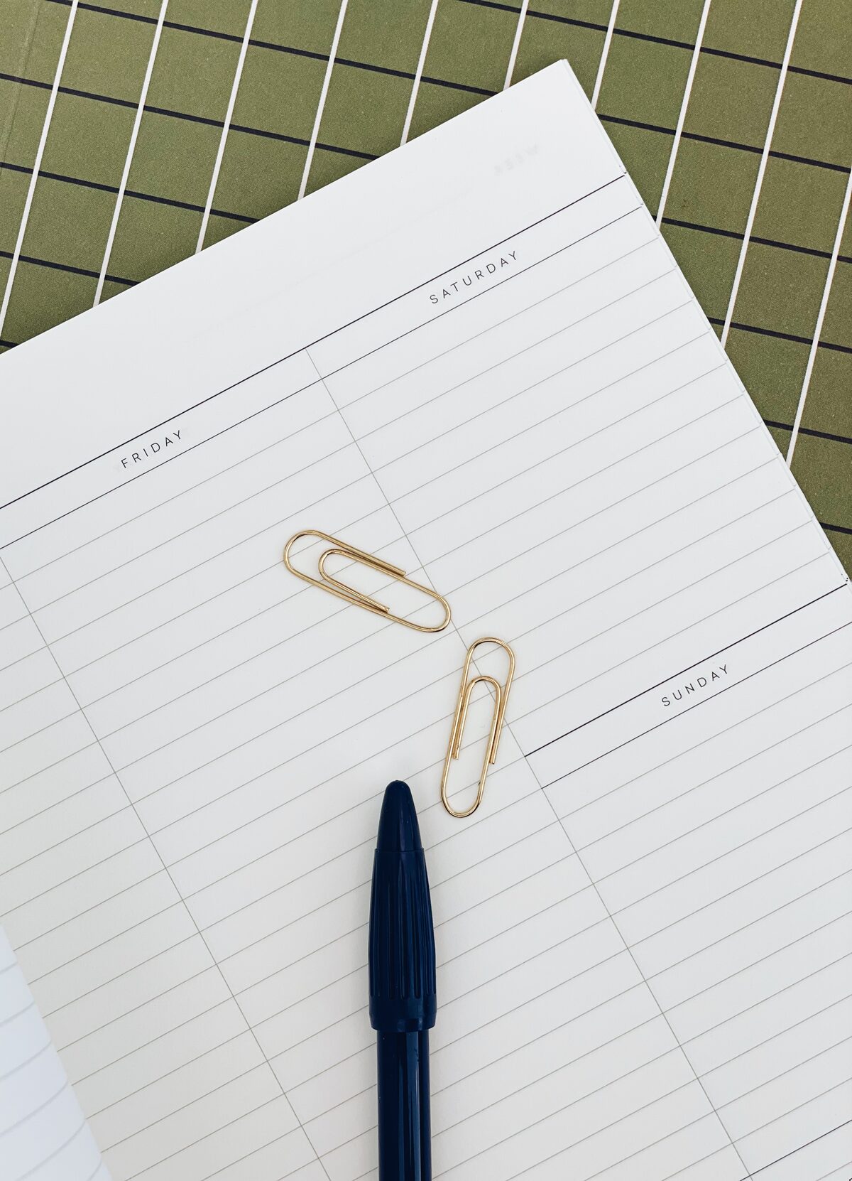 A top-down view of a lined weekly planner page with gold paperclips and a dark blue pen on a green-and-white checkered surface.