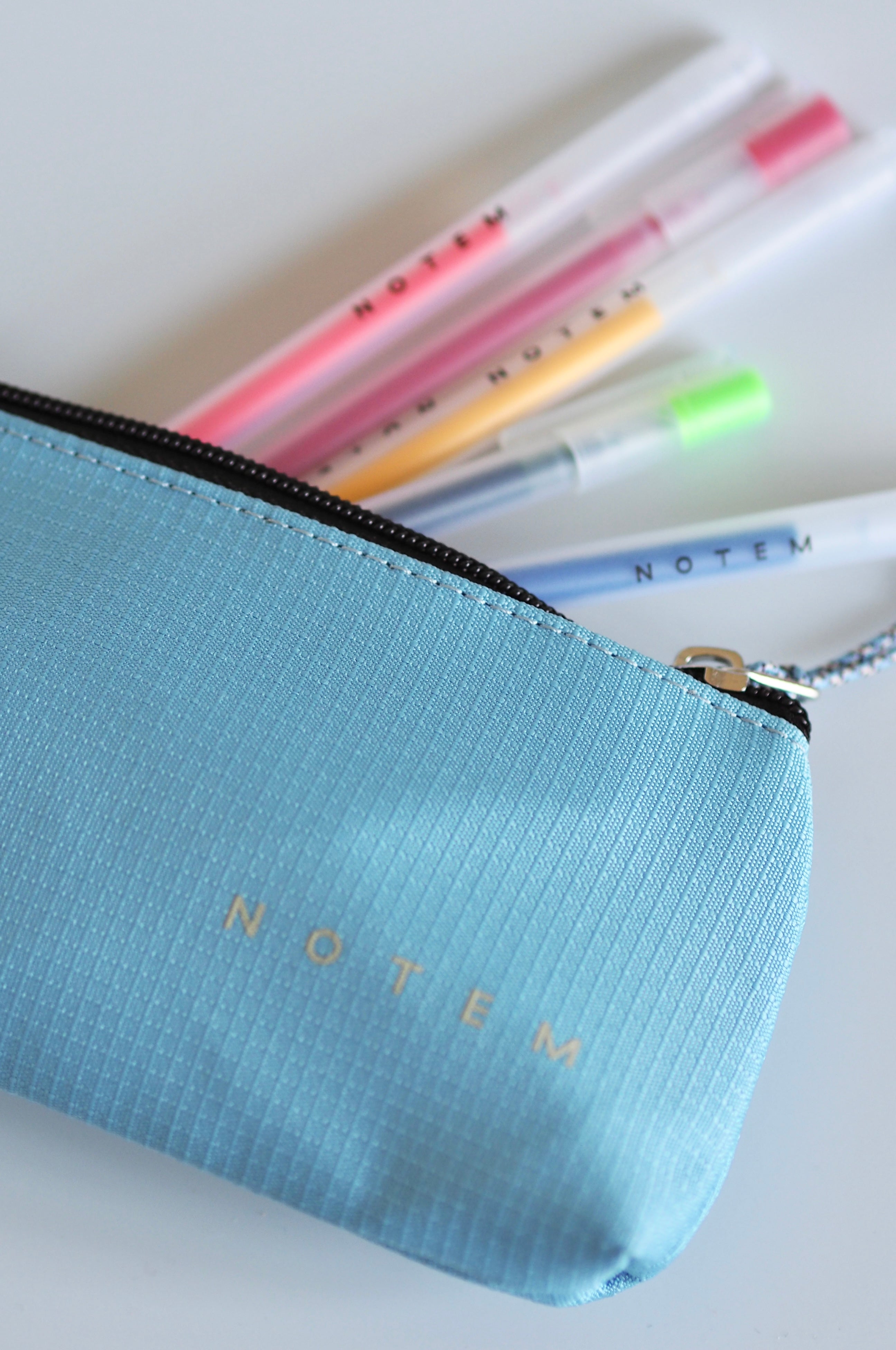 A light blue textured zippered pouch with the word 'NOTEM' printed in gold on the front, shown with several pastel highlighters behind it on a white background.